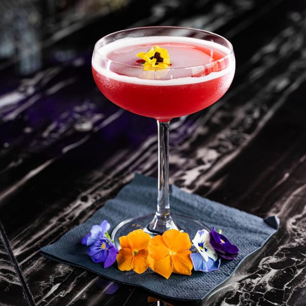 A vibrant red cocktail with a yellow edible flower, surrounded by colorful petals on a dark napkin.