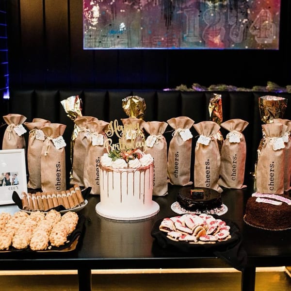 A dessert table with cakes, cookies, and gift bags, set against a dark background with abstract art.