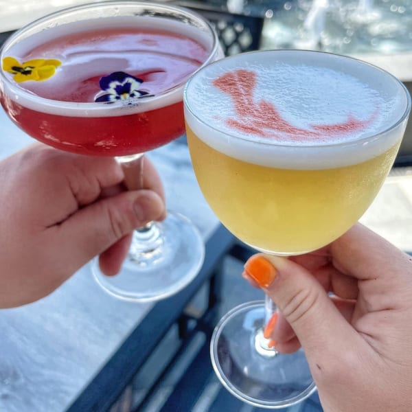 Two hands holding colorful cocktails, one red with flowers, one yellow with a red design.